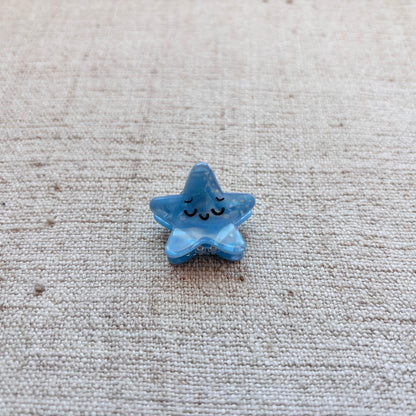 A small light blue star-shaped hair clip with a simple black line face, placed on a textured beige fabric background.