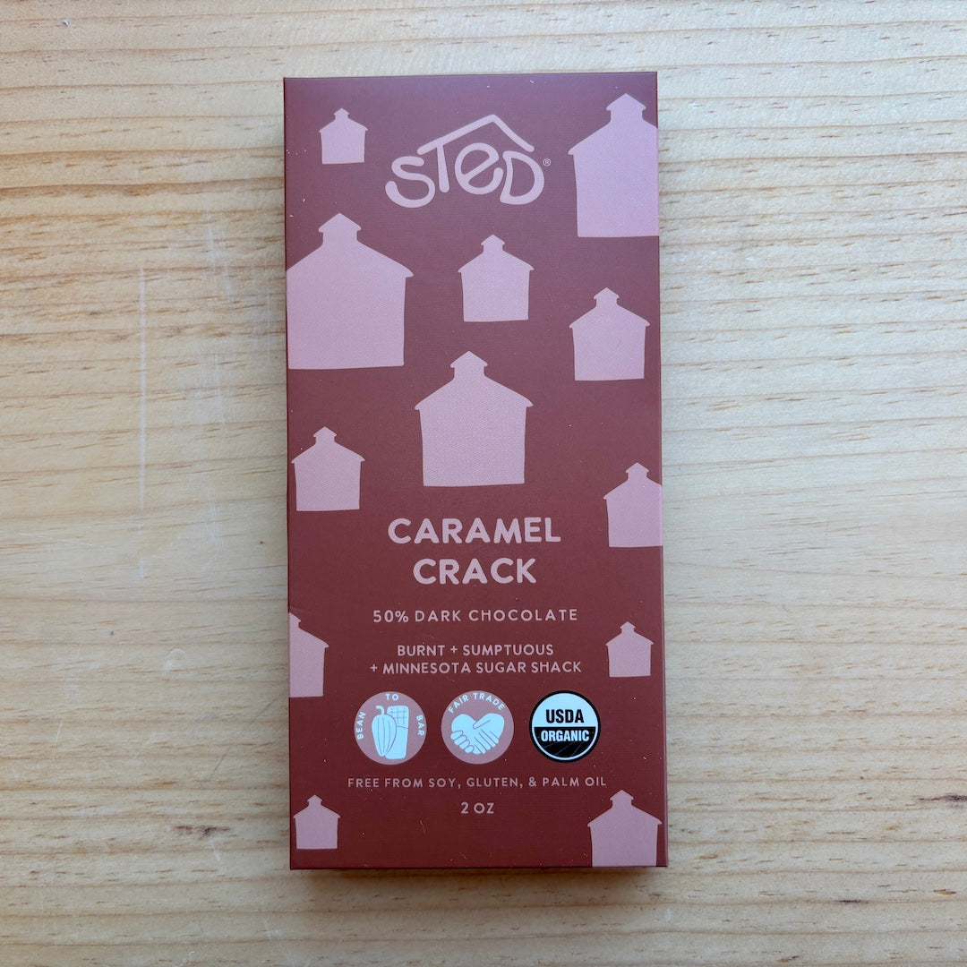 A 2 oz bar of Sted Caramel Crack chocolate, featuring a dark reddish-brown package with light pink house silhouettes and white text, placed on a light wood background.