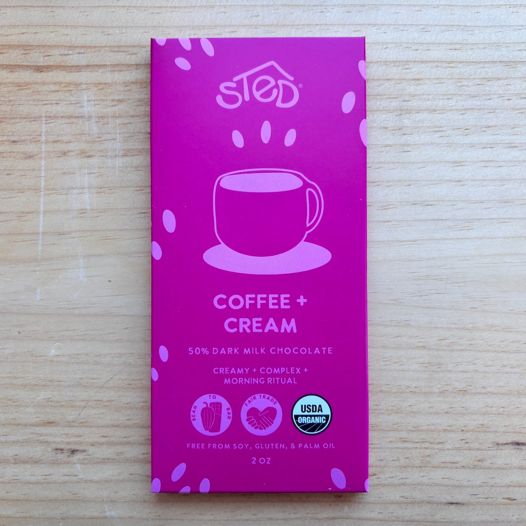 A 2 oz bar of sted Coffee + Cream chocolate in its pink packaging, placed flat on a light wood surface.