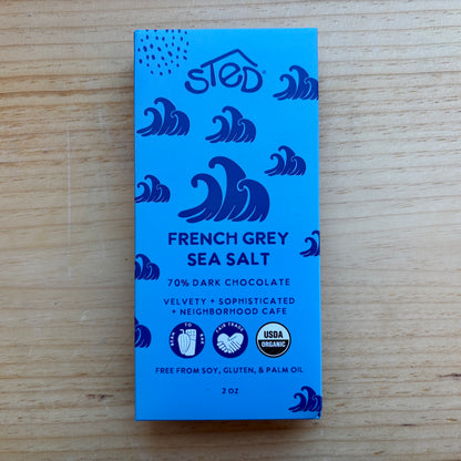 A blue chocolate bar package labeled 'sted' and 'French Grey Sea Salt' with dark blue wave illustrations, photographed on a light wood surface.