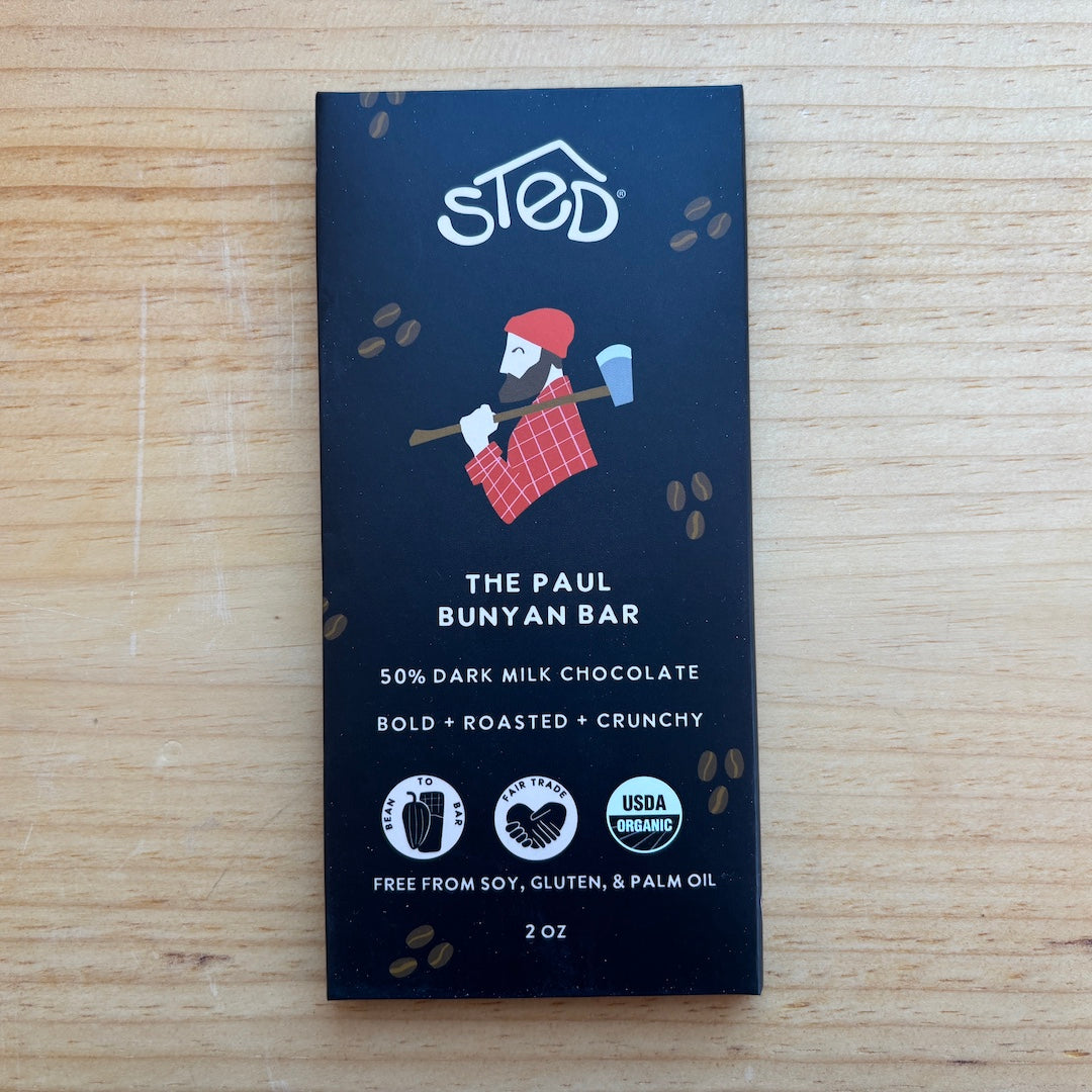A 2 oz bar of 'The Paul' Bunyan Bar chocolate by 'sted.' in its dark navy blue packaging, placed flat on a light wood grain surface.