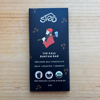 A 2 oz bar of 'The Paul' Bunyan Bar chocolate by 'sted.' in its dark navy blue packaging, placed flat on a light wood grain surface.
