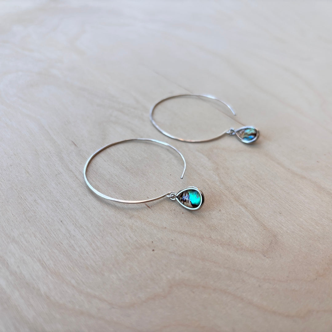 Silver hoop earrings with teardrop-shaped paua dangle on a light wooden surface