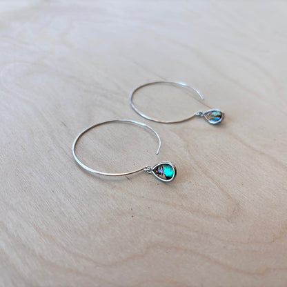 Silver hoop earrings with teardrop-shaped paua dangle on a light wooden surface