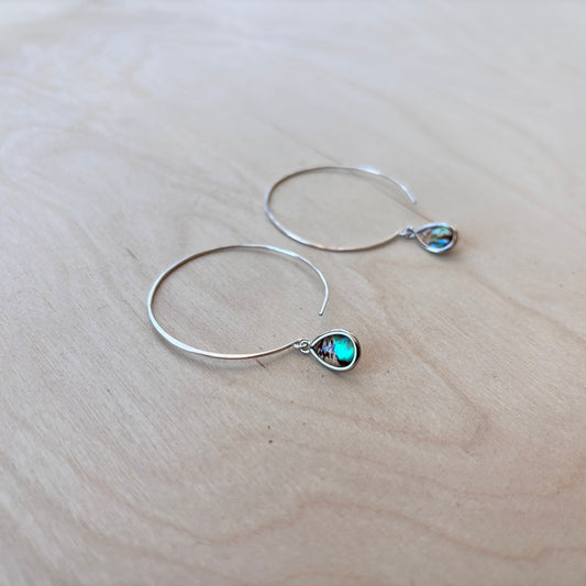 Silver hoop earrings with teardrop-shaped paua dangle on a light wooden surface