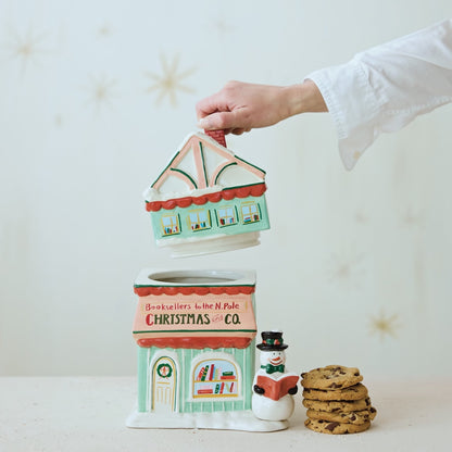 A ceramic Christmas-themed cookie jar shaped like a storefront, with a hand lifting the lid to reveal a cookie compartment. A stack of cookies sits beside it on a light surface against a soft cream background with faint star decorations.