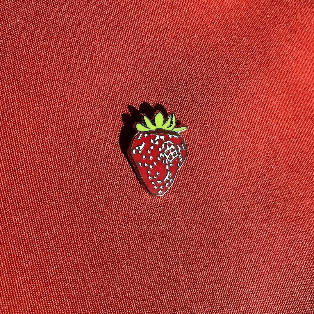 A strawberry-shaped enamel pin with a red body, white seeds, and green leafy top, placed on a textured red fabric background.