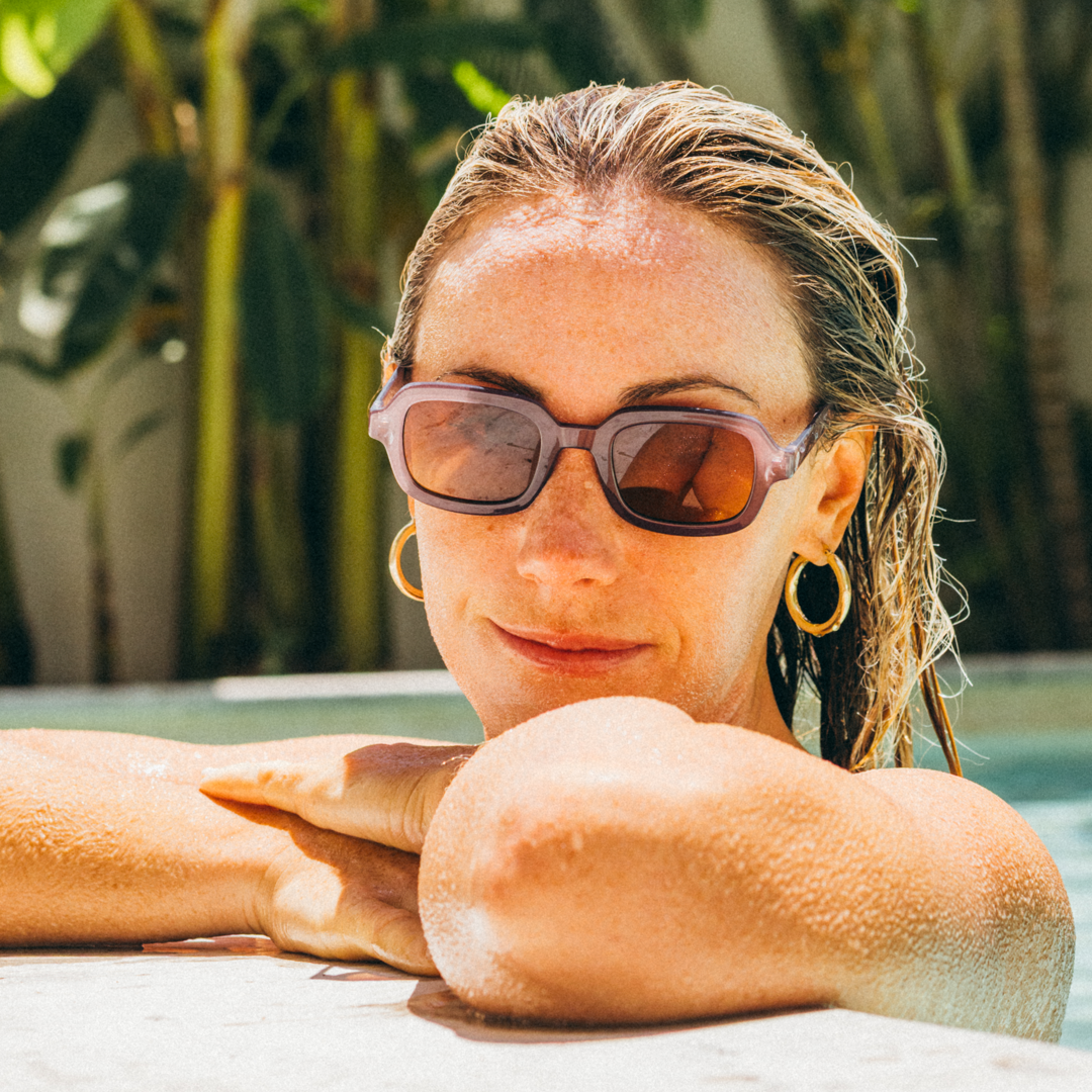 A woman with wet hair leans on a poolside, wearing purple-framed sunglasses with dark brown lenses and gold hoop earrings.