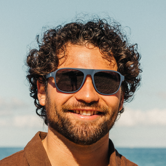 A close-up portrait of a smiling man with curly dark hair and a beard, wearing dark sunglasses with a blue frame, set against a soft outdoor background of sky and water.