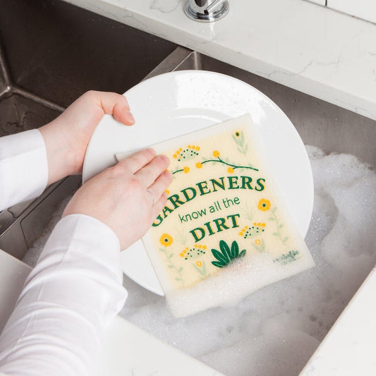 Hand washing a dish with a Swedish dishcloth featuring a gardening-themed message.