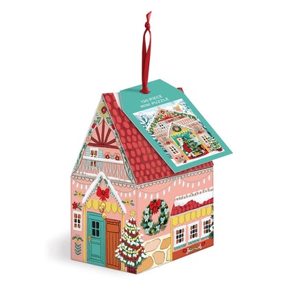A Christmas-themed jigsaw puzzle box shaped like a gingerbread house, featuring a red hanging ribbon and a teal tag that reads '130-piece mini puzzle'. The box is decorated with festive illustrations including a decorated Christmas tree, wreaths, and a red patterned roof, set against a plain white background.