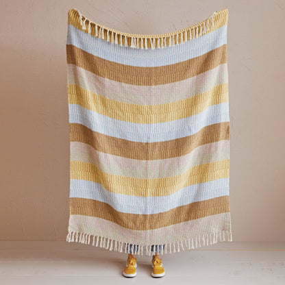 A striped knit blanket with fringe on both ends is held up against a plain beige wall. The blanket has horizontal stripes in tan, light blue, and gold.