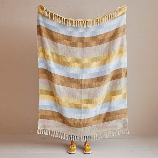 A striped knit blanket with fringe on both ends is held up against a plain beige wall. The blanket has horizontal stripes in tan, light blue, and gold.