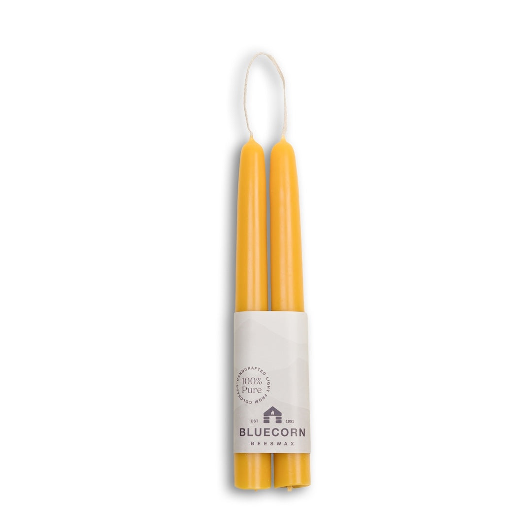 Two yellow taper candles in a double-wrapped package with a white paper band labeled 'BlueCorn Beeswax' and '100% Pure'.