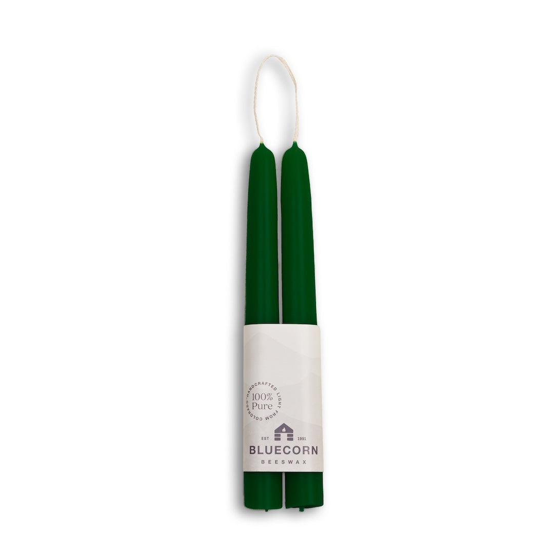 A pair of dark green taper candles wrapped in a white paper band labeled 'BLUECORN Beeswax', shown against a plain white background.