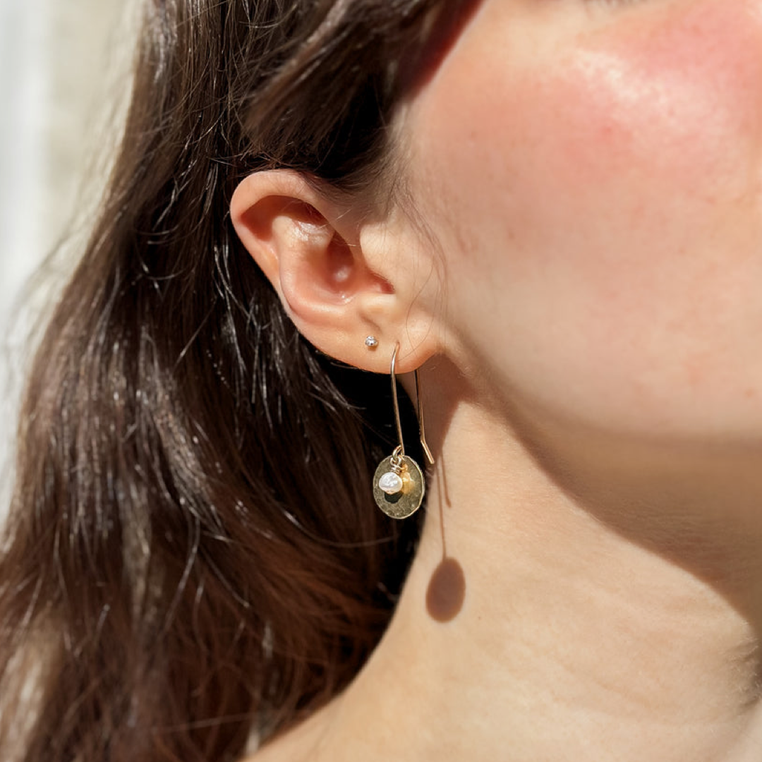 A close-up lifestyle shot of a woman's ear wearing a delicate gold dangle earring with a small round gold charm and a tiny freshwater pearl, set against her cheek and dark hair.