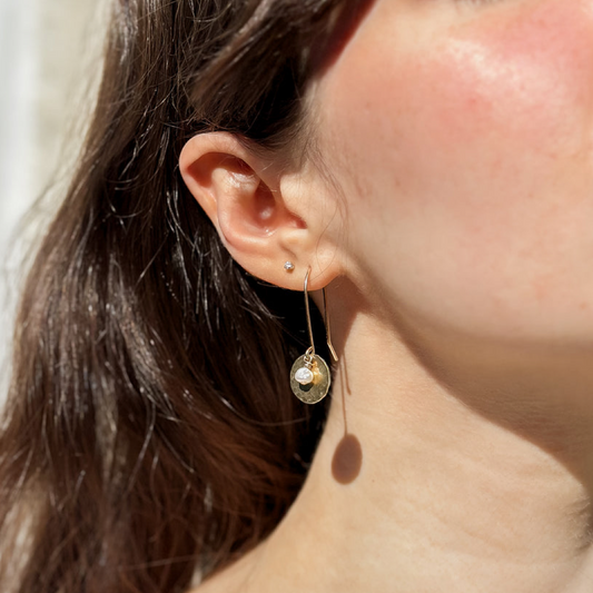 A close-up lifestyle shot of a woman's ear wearing a delicate gold dangle earring with a small round gold charm and a tiny freshwater pearl, set against her cheek and dark hair.
