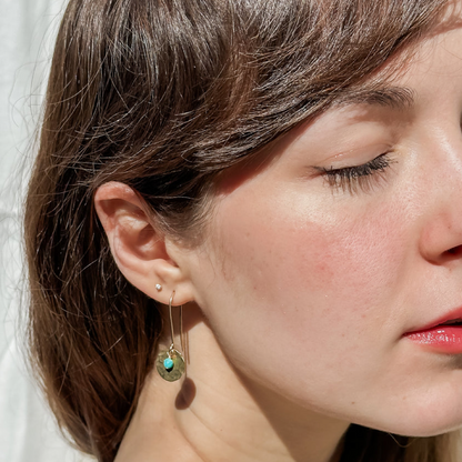 A close-up lifestyle shot of a woman's ear wearing a delicate gold dangle earring with a small round gold charm and a tiny turquoise gemstone set against her cheek and dark hair.