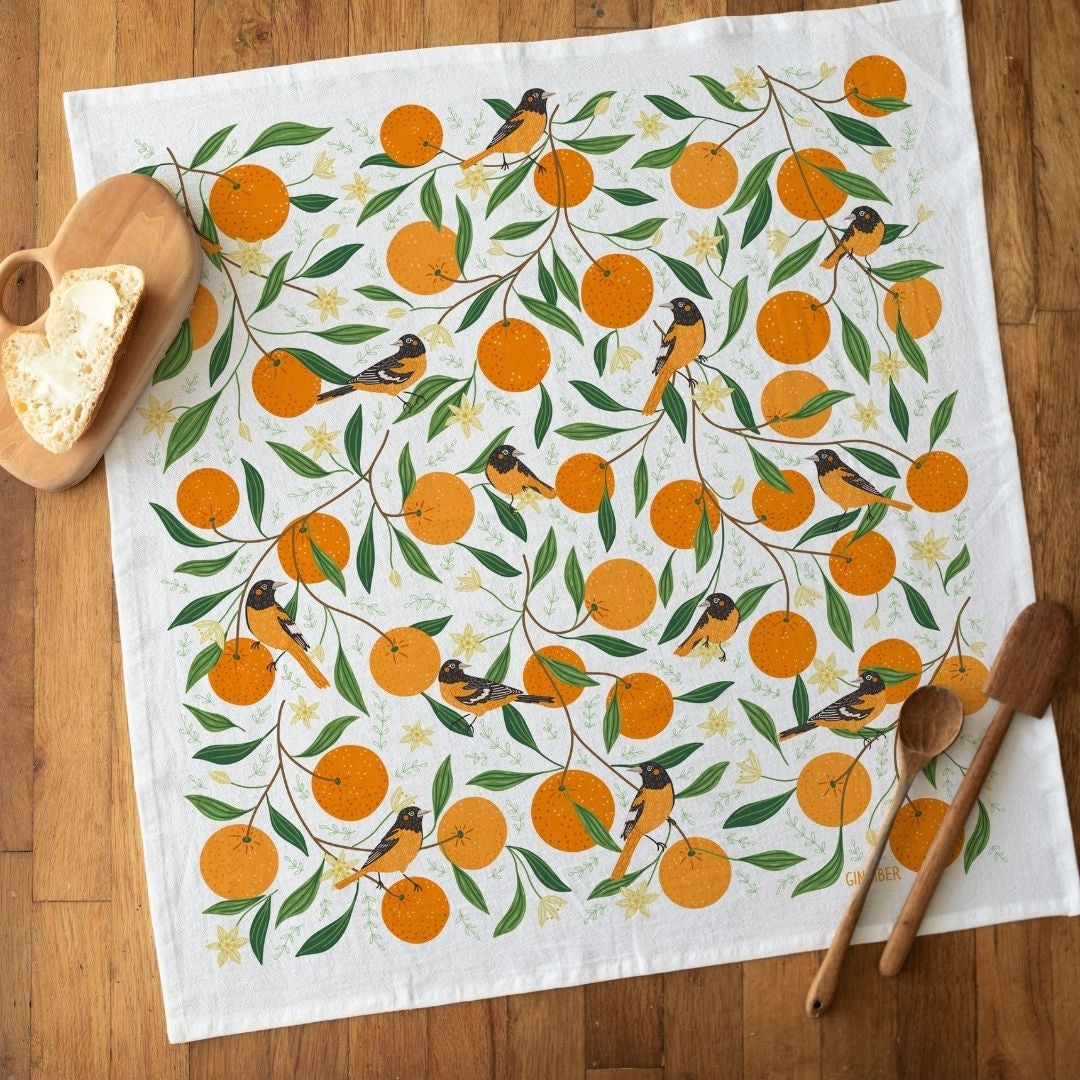 A square white tea towel laid flat on a wooden table, printed with an allover pattern of orange oranges, green leaves, and small birds. A wooden spoon, a wooden spoon handle, and a wooden heart-shaped object with a slice of bread are placed around it as styling props.