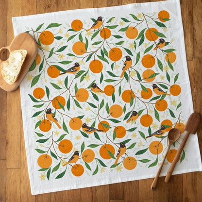 A square white tea towel laid flat on a wooden table, printed with an allover pattern of orange oranges, green leaves, and small birds. A wooden spoon, a wooden spoon handle, and a wooden heart-shaped object with a slice of bread are placed around it as styling props.