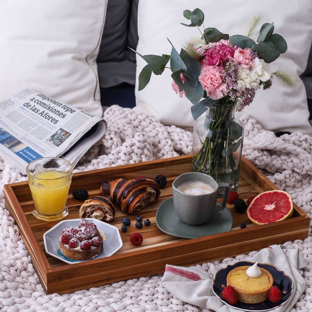 Wooden tray with breakfast items including coffee, pastries, and fruit on a cozy blanket.