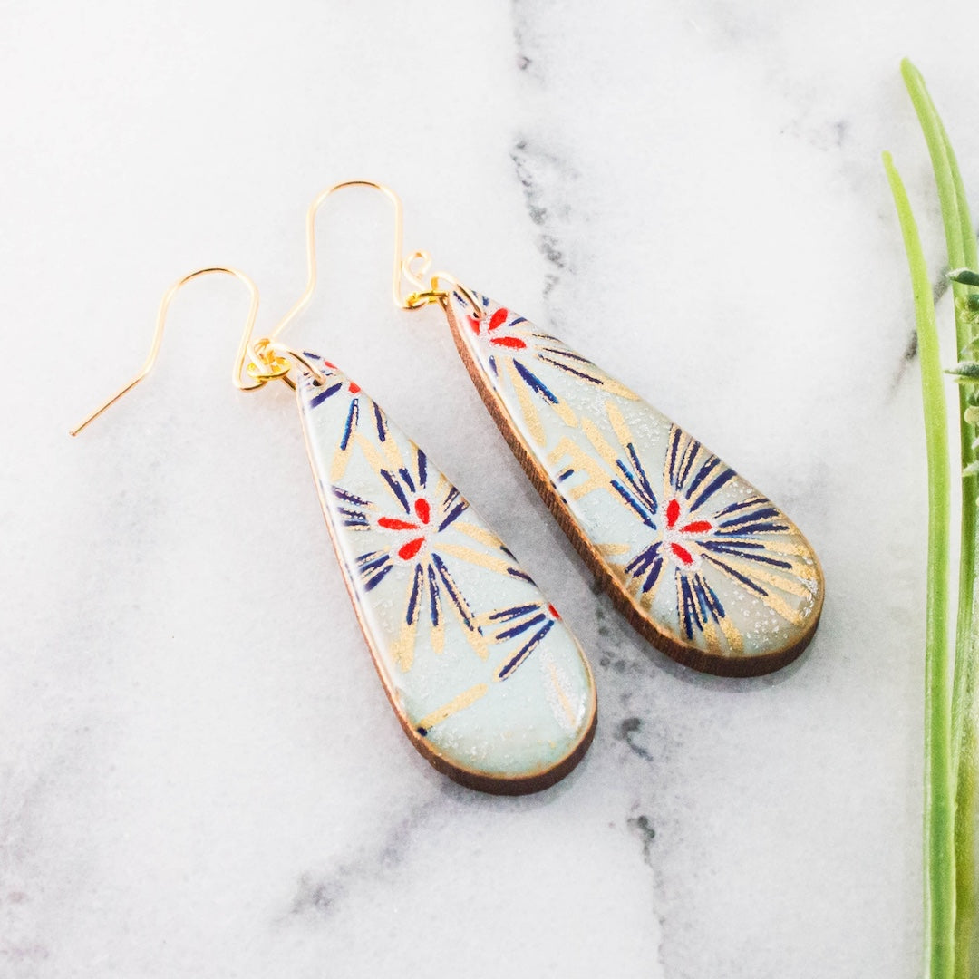 A pair of teardrop-shaped dangling earrings with gold-tone wires and a light blue-gray base, decorated with navy and red starburst patterns and gold radial lines, displayed on a white marble surface.
