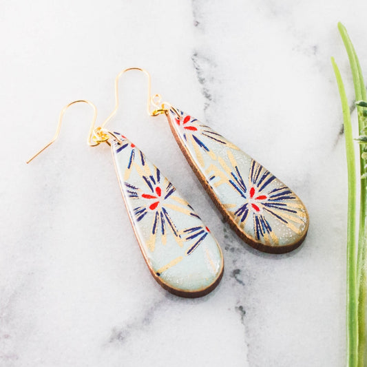 A pair of teardrop-shaped dangling earrings with gold-tone wires and a light blue-gray base, decorated with navy and red starburst patterns and gold radial lines, displayed on a white marble surface.