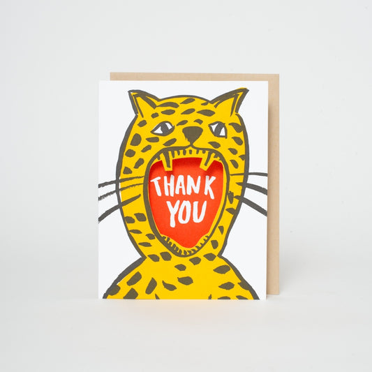 A greeting card featuring a cartoon cheetah with its mouth wide open, revealing the words 'THANK YOU' in white on a red background. The card is shown standing upright next to a light brown envelope against a clean white background.