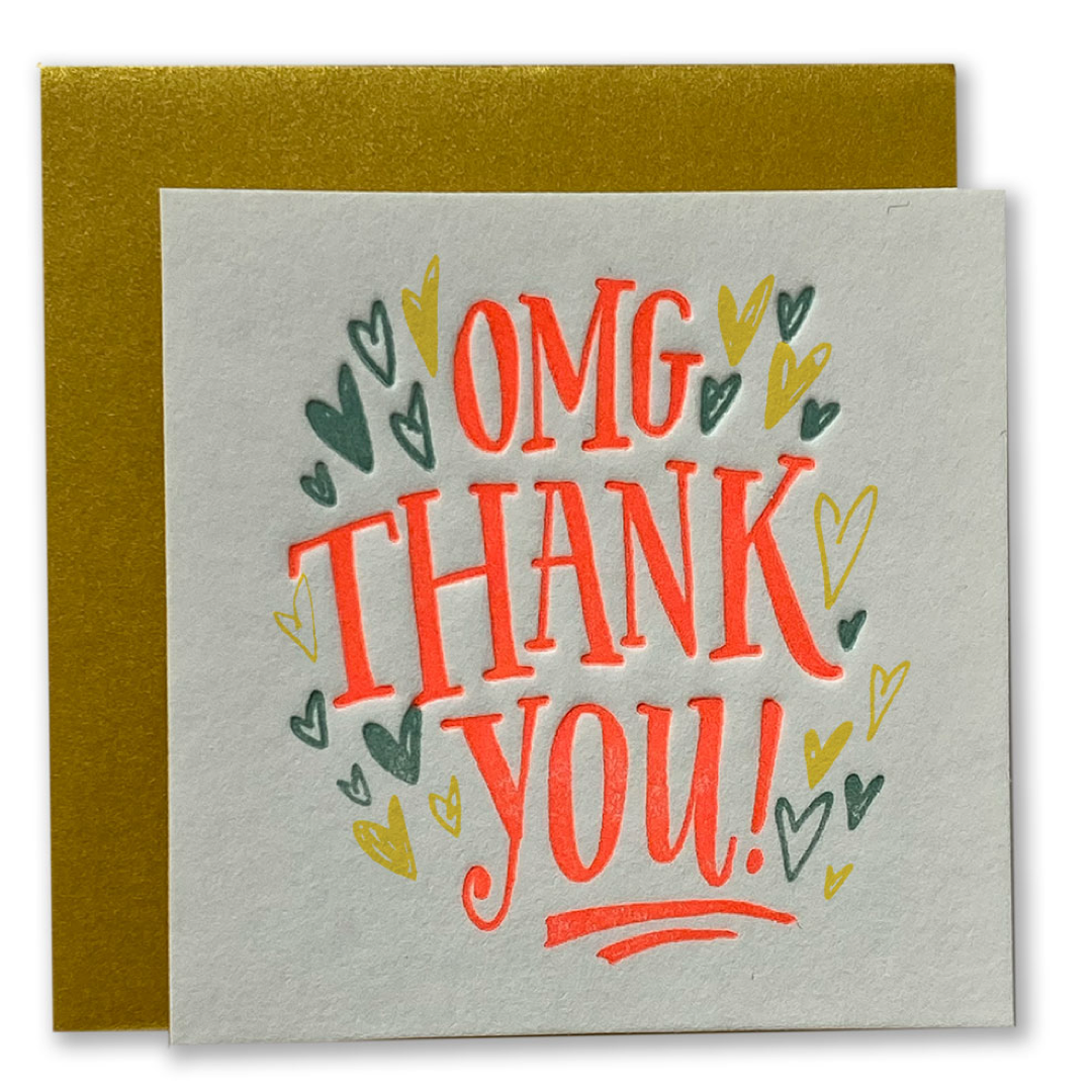 A square greeting card with a textured off-white paper front and a metallic gold envelope behind it. The card features the text 'OMG THANK YOU!' in a playful orange-red font, surrounded by small hand-drawn hearts in orange, yellow, and dark green.