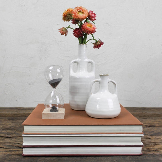 Stack of books with a vase, hourglass, and flowers on top against a white brick wall.