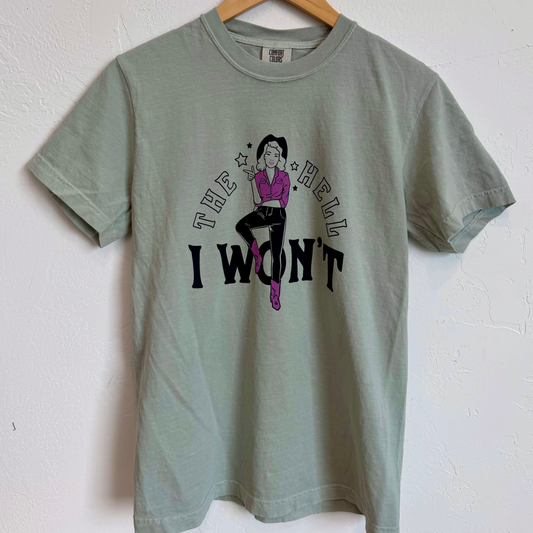 A light sage green graphic t-shirt hanging on a wooden hanger against a white textured wall. The shirt features a graphic of a woman in a purple top and pink boots with the text 'THE HELL I WON'T' in black.