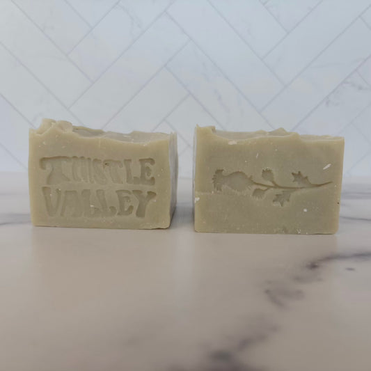 Two handmade soap bars in a natural light green color, displayed side-by-side on a marble surface with a white tiled backsplash. The left bar is embossed with the words 'THISTLE VALLEY', and the right bar features a stamped design of a thistle