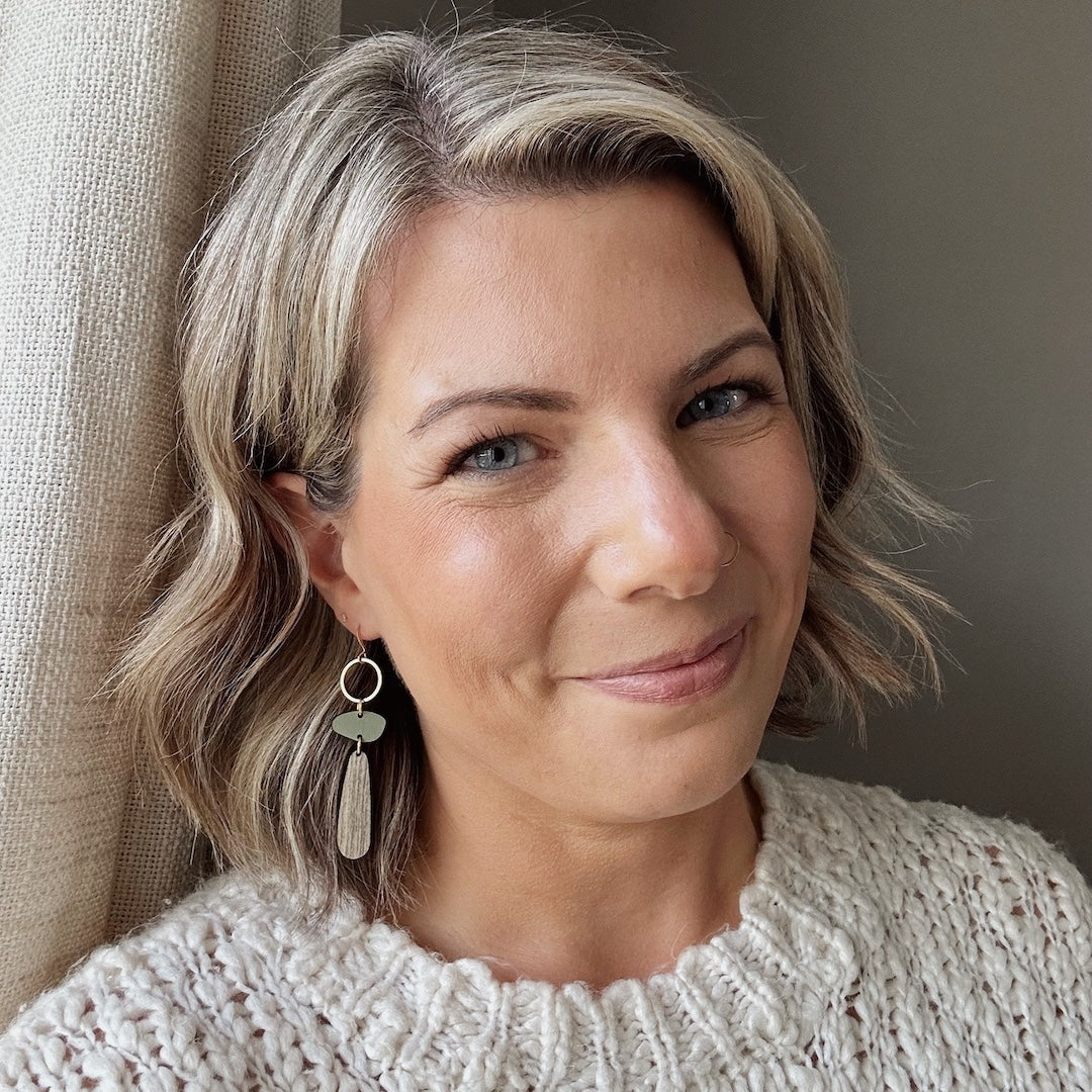Woman with short blonde hair wearing a beige sweater and earrings, standing against a neutral background.
