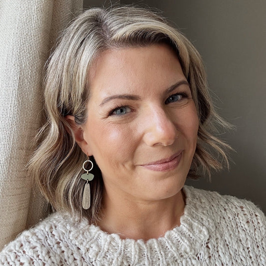 Woman with short blonde hair wearing a beige sweater and earrings, standing against a neutral background.