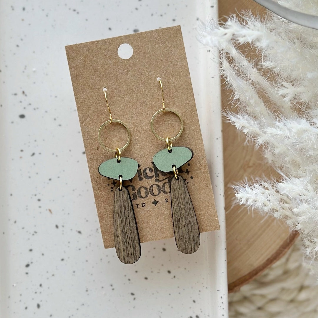 Pair of earrings on a cardboard backing with a textured white background. The earrings are long dangle earrings that are made of wood and meant to resemble a tree