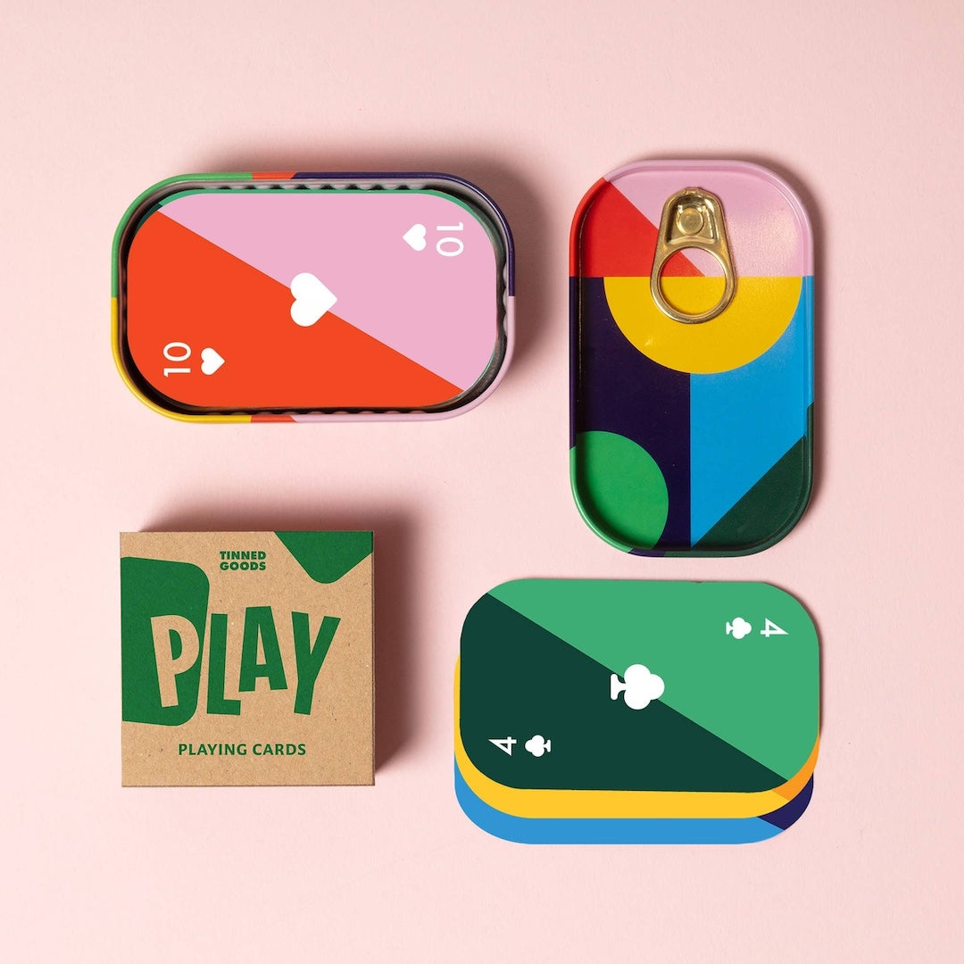 A set of colorful playing cards and their packaging arranged on a light pink background. The cards are designed to look like sardine tins with abstract geometric color blocks and playing card suits.