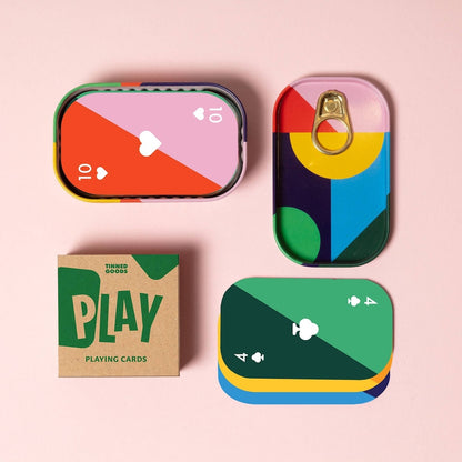 A set of colorful playing cards and their packaging arranged on a light pink background. The cards are designed to look like sardine tins with abstract geometric color blocks and playing card suits.