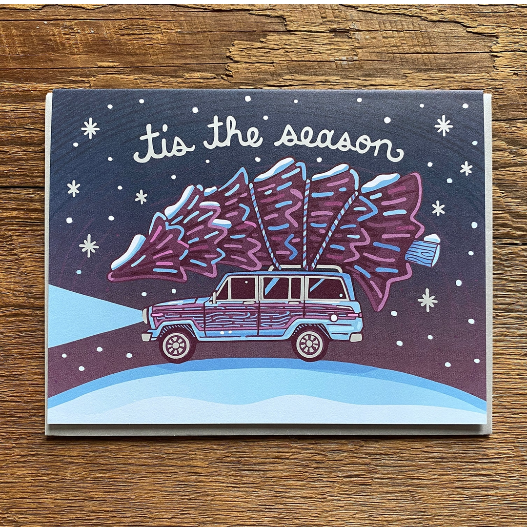 A square Christmas greeting card featuring an illustrated purple and blue station wagon carrying a Christmas tree on its roof, with the text 'It's the season' on a snowy night scene, photographed on a wooden surface.