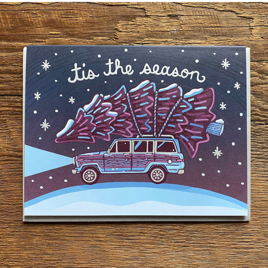 A square Christmas greeting card featuring an illustrated purple and blue station wagon carrying a Christmas tree on its roof, with the text 'It's the season' on a snowy night scene, photographed on a wooden surface.