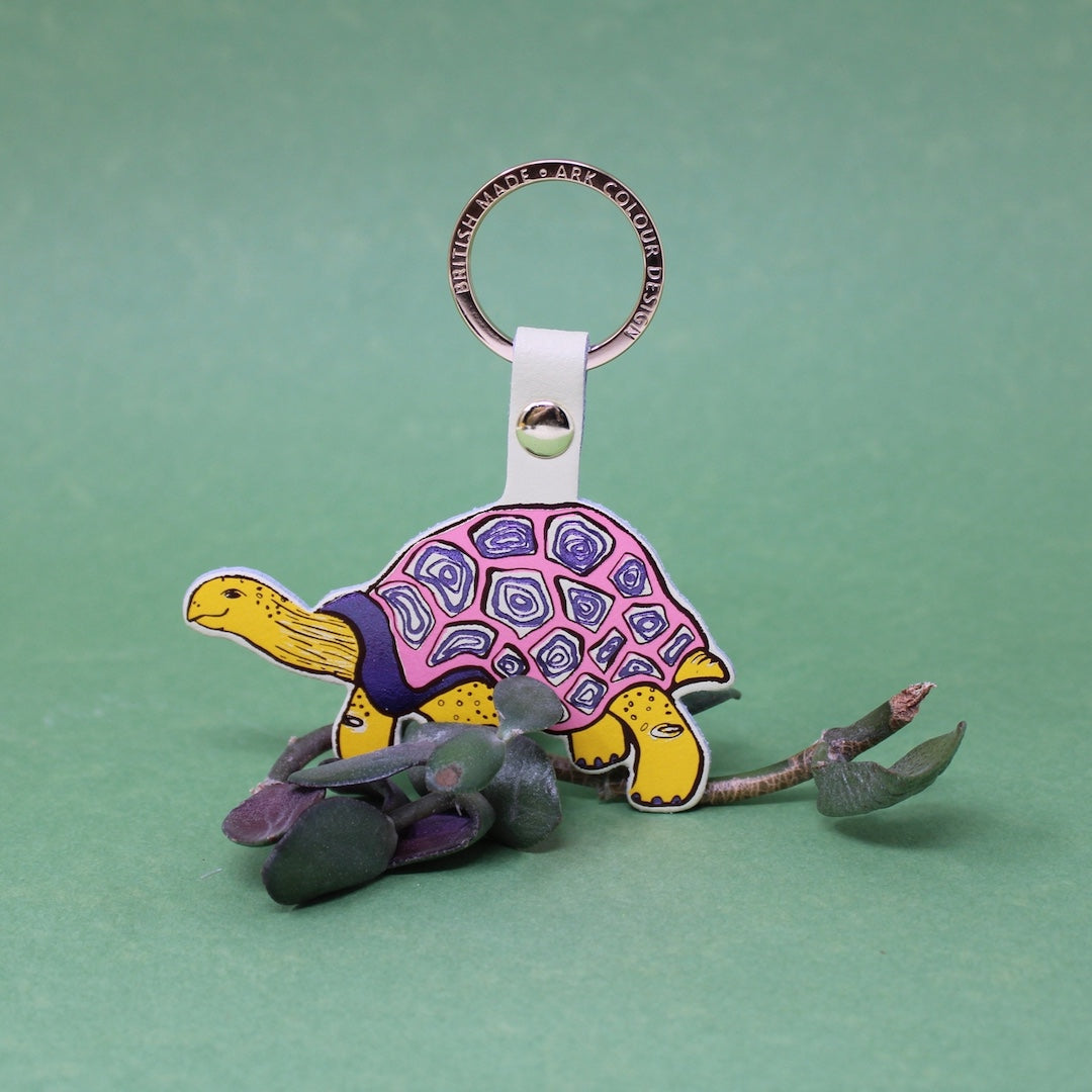 A colorful turtle-shaped keychain with a pink and purple patterned shell, yellow head and legs, and gray accents, attached to a metal key ring on a solid green background.