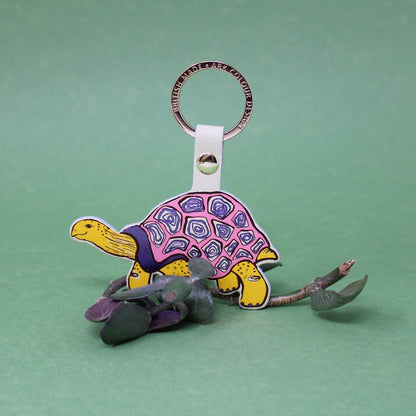 A colorful turtle-shaped keychain with a pink and purple patterned shell, yellow head and legs, and gray accents, attached to a metal key ring on a solid green background.