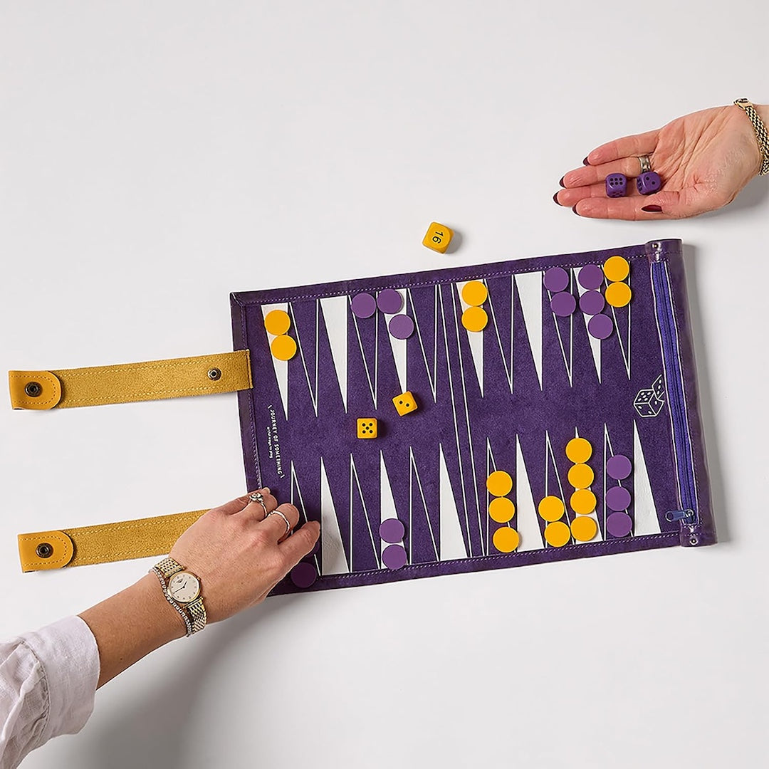 A purple backgammon board with yellow straps and white playing points, shown on a light gray background with two hands holding it open and yellow and purple dice scattered around.