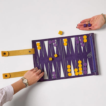 A purple backgammon board with yellow straps and white playing points, shown on a light gray background with two hands holding it open and yellow and purple dice scattered around.