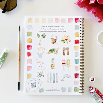 Artistic coloring book with watercolor tropical vacation themed illustrations on a white surface with art supplies.