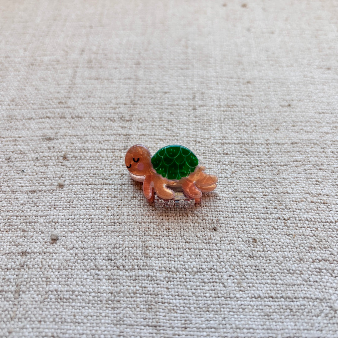 A small turtle-shaped hair clip with a brown body and a green patterned shell, resting on a textured beige fabric background.