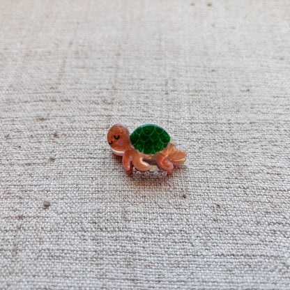 A small turtle-shaped hair clip with a brown body and a green patterned shell, resting on a textured beige fabric background.
