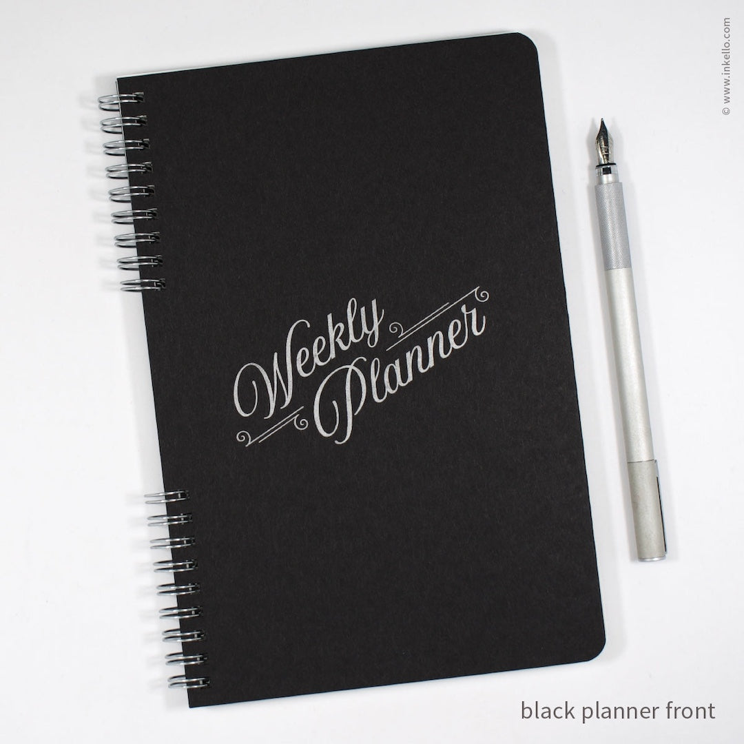 A black spiral-bound weekly planner with silver lettering on the cover, shown on a white background next to a silver pen. Text in the corner reads 'black planner front'.