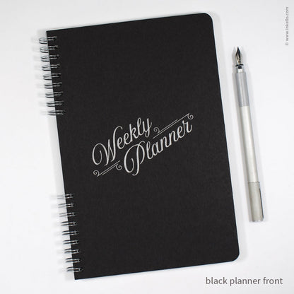 A black spiral-bound weekly planner with silver lettering on the cover, shown on a white background next to a silver pen. Text in the corner reads 'black planner front'.
