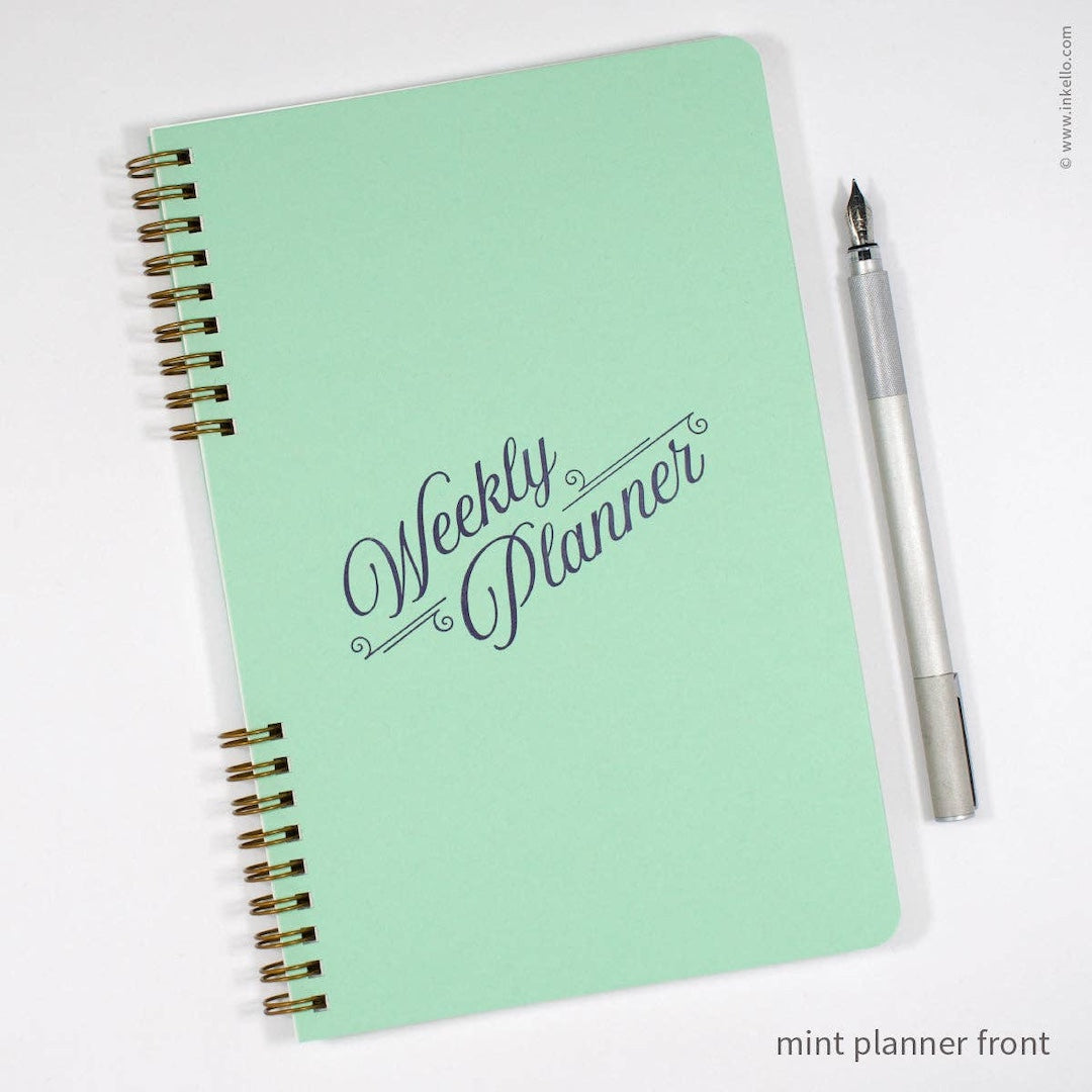 A mint green weekly planner notebook with gold spiral binding and 'Weekly Planner' written in dark gray script on the cover, shown on a white background next to a silver pen.