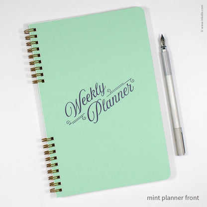 A mint green weekly planner notebook with gold spiral binding and 'Weekly Planner' written in dark gray script on the cover, shown on a white background next to a silver pen.